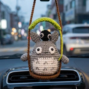 Puede incluir: Amuleto de coche de ganchillo con un personaje gris parecido a Totoro con un duende de hollín negro encima, suspendido de un columpio marrón y verde. El amuleto cuelga del espejo retrovisor dentro de un coche, con una escena de calle borrosa en el fondo.