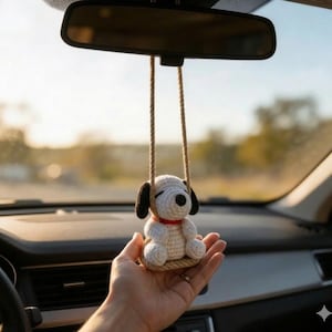 K&ouml;nnte beinhalten: Ein geh&auml;kelter Hunde-Autoanh&auml;nger in Wei&szlig; und Schwarz mit rotem Halsband, der an einem R&uuml;ckspiegel h&auml;ngt. Der Anh&auml;nger wird von zwei beigefarbenen Kordeln gehalten. Der Anh&auml;nger wird in der Hand einer Person gehalten.