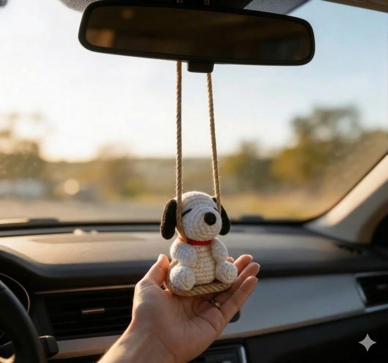K&ouml;nnte beinhalten: Ein geh&auml;kelter Hunde-Autoanh&auml;nger in Wei&szlig; und Schwarz mit rotem Halsband, der an einem R&uuml;ckspiegel h&auml;ngt. Der Anh&auml;nger wird von zwei beigefarbenen Kordeln gehalten. Der Anh&auml;nger wird in der Hand einer Person gehalten.