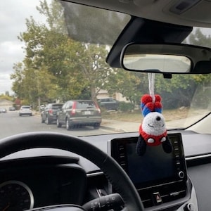 K&ouml;nnte beinhalten: Innenansicht eines Autos mit schwarzem Lenkrad und Armaturenbrett. Ein handgefertigter Panda-Autoanh&auml;nger h&auml;ngt am R&uuml;ckspiegel. Der Panda tr&auml;gt ein rot-blaues Outfit mit schwarzen Akzenten und einem l&auml;chelnden Gesicht. Autos und B&auml;ume s&auml;umen die Stra&szlig;e drau&szlig;en.