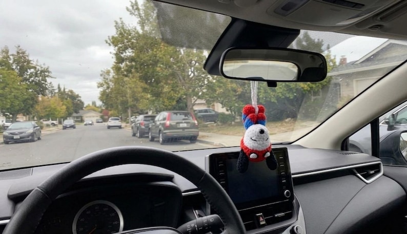 K&ouml;nnte beinhalten: Innenansicht eines Autos mit schwarzem Lenkrad und Armaturenbrett. Ein handgefertigter Panda-Autoanh&auml;nger h&auml;ngt am R&uuml;ckspiegel. Der Panda tr&auml;gt ein rot-blaues Outfit mit schwarzen Akzenten und einem l&auml;chelnden Gesicht. Autos und B&auml;ume s&auml;umen die Stra&szlig;e drau&szlig;en.