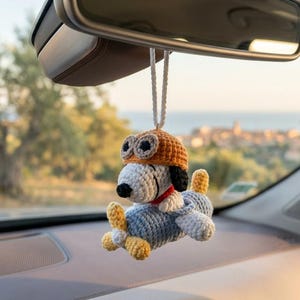Può includere: Ciondolo per auto di Snoopy all'uncinetto, raffigurante Snoopy come pilota con cappello arancione e aereo. Il ciondolo è bianco, nero, blu, arancione e giallo. È appeso a un filo bianco, sospeso dallo specchietto retrovisore di un'auto.