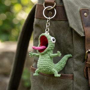 May include: A green crochet lizard keyring with a wide open red mouth and pink tongue. The keyring is attached to a silver ring and hangs from a green canvas rucksack with brown leather accents.