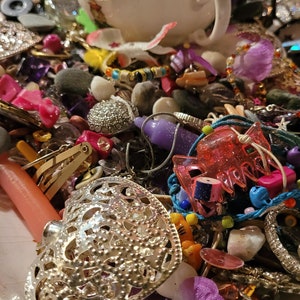 May include: A white teacup rests amidst a collection of jewellery, beads, and trinkets. A silver filigree heart-shaped pendant is in the foreground, surrounded by pink, purple, and clear beads. The items are on a white plate with a green floral design.