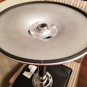 Op de afbeelding: Een ronde, zilverkleurige bartafel met een zwarte rand. De tafel heeft een chroomkleurige voet en een zwarte zitting. De diameter van de tafel is ongeveer 61 cm.
