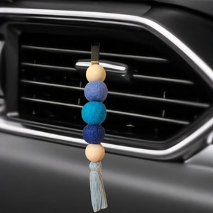 May include: A blue and beige car air freshener with a tassel. The air freshener is made of felt balls and wooden beads.