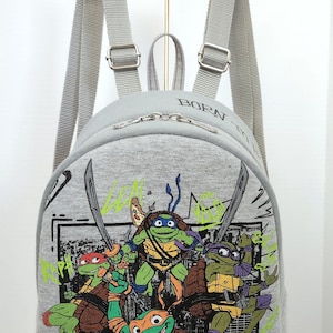 May include: A gray backpack featuring the Teenage Mutant Ninja Turtles. The front of the backpack displays the four turtles with their signature weapons and colors. The word "BORN" is printed on the top of the backpack.