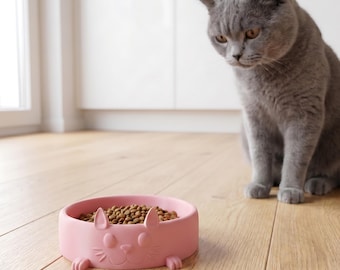 May include: A round, light pink pet bowl featuring a molded cat face design on the front, filled with brown dry kibble. A grey cat sits nearby, looking down at the bowl. The text "Shapid" appears in a yellow circle in the bottom right corner.