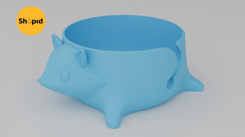 May include: A light blue yarn bowl shaped like a stylized hedgehog with a smooth matte finish. The bowl features a circular opening at the top and a J-shaped cutout on the side for yarn to pass through. A yellow circular logo with the text Shpid is visible.