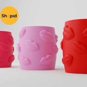 May include: Three cylindrical cups with a lip print design. The cups are red, pink, and light pink.
