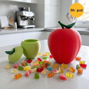 Peut inclure: Un récipient rouge en forme de pomme avec une tige verte et un récipient vert assorti avec un couvercle. Des bonbons gélifiés colorés sont dispersés autour des récipients. Les récipients sont sur un comptoir blanc.