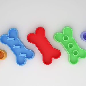 May include: Five dog bone shaped slow feeder bowls in different colors: orange, blue, red, green, and purple. Each bowl has a different design of raised bumps and ridges to slow down eating.
