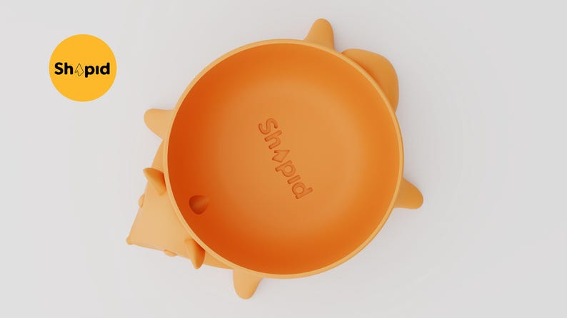 May include: An orange silicone bowl shaped like a hedgehog, featuring the embossed text "Shapid" in the center. The bowl has small, rounded protrusions along the sides representing the hedgehog's spikes, and a small circular hole near the bottom rim.