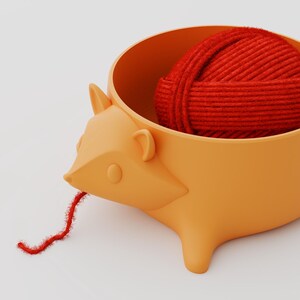May include: A light orange hedgehog-shaped yarn bowl with a smooth matte finish. Inside the bowl sits a round ball of vibrant red yarn, with a single strand of the red yarn extending out from the hedgehog's mouth. The text Shpid is visible.