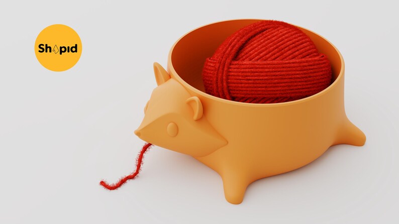 May include: A light orange hedgehog-shaped yarn bowl with a smooth matte finish. Inside the bowl sits a round ball of vibrant red yarn, with a single strand of the red yarn extending out from the hedgehog's mouth. The text Shpid is visible.