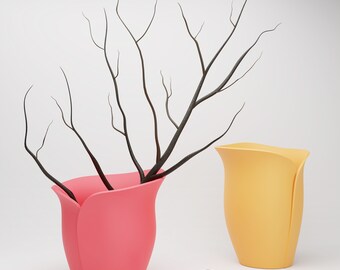 May include: Two flower-shaped vases, one pink and one yellow, with a dried branch in the pink vase.  There are also a few pink flower petals on the white surface.