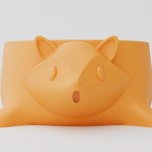 May include: A matte orange planter shaped like a tree stump with a stylized fox face embossed on the front. The fox features pointed ears, oval eyes, and a small open mouth. A yellow circular logo with the text Shapid is visible in the top left.