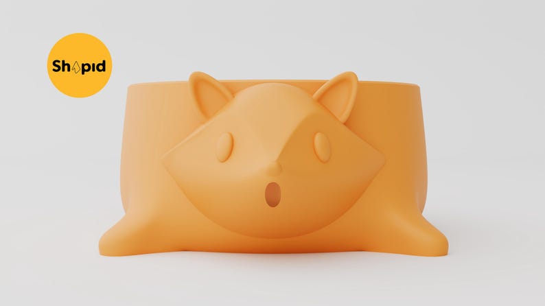 May include: A matte orange planter shaped like a tree stump with a stylized fox face embossed on the front. The fox features pointed ears, oval eyes, and a small open mouth. A yellow circular logo with the text Shapid is visible in the top left.