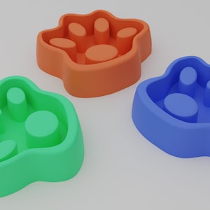 May include: Three paw-shaped slow feeder bowls for pets. The bowls are made of silicone and come in green, blue, and orange.