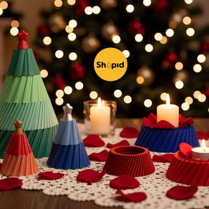 May include: A festive Christmas display featuring decorative paper Christmas trees and candle holders in various colors. Lit candles and red rose petals are scattered on a lace tablecloth, with a Christmas tree in the background.