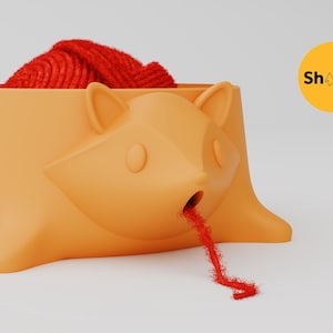 May include: An orange yarn bowl shaped like a stylized fox head with pointed ears and a small circular opening at the mouth. A ball of red yarn sits inside the bowl, with a single strand of red yarn threaded through the fox's mouth. Text reads Shapid.