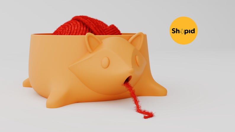 May include: An orange yarn bowl shaped like a stylized fox head with pointed ears and a small circular opening at the mouth. A ball of red yarn sits inside the bowl, with a single strand of red yarn threaded through the fox's mouth. Text reads Shapid.