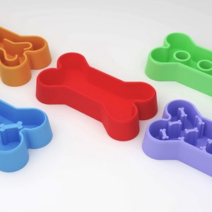 Op de afbeelding: Vijf hondenbakken in de vorm van een bot in verschillende kleuren: oranje, rood, groen, blauw en paars. De bakken hebben verschillende ontwerpen op de bodem om het eten te vertragen.