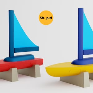 May include: Two toy sailboats, one red and one yellow, each with a blue sail and mast. The boats are mounted on beige supports. The word "Shopid" is in a yellow circle in the background.