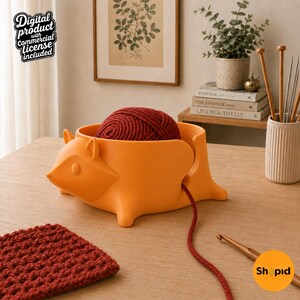May include: An orange yarn bowl shaped like a stylized fox, containing a ball of dark red yarn. A single strand of the red yarn extends from the bowl through a side opening. The bowl features a fox head on one side and four small feet at the base.