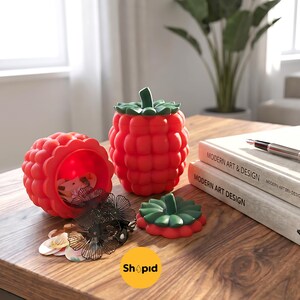 Puede incluir: Un recipiente rojo con forma de frambuesa con un tallo y una tapa verdes, junto a un recipiente abierto y una tapa. El recipiente está sobre una superficie de madera con accesorios para el cabello y libros. La imagen incluye el texto "Shopid".