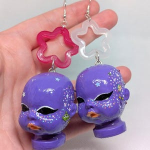 Op de afbeelding: Een paar bungelende oorbellen met paarse poppenhoofdjes, beschilderd met zwarte eyeliner en oranje lippen. Elk hoofdje is versierd met kleine, kleurrijke ruimtevaartstickers en glitters. De oorbellen hangen aan zilverkleurige haakjes met doorschijnende roze en witte stervormige bedels.