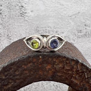 May include: A silver ring featuring an eye design. Each eye has a bezel-set gemstone: a round, light green peridot and a round, dark blue iolite. The ring is set on a textured, dark brown surface.