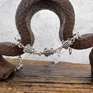 May include: A silver bracelet with five turtle charms. The bracelet is shown against a rustic wooden surface and a large, textured metal object. The turtles are linked by a delicate chain.