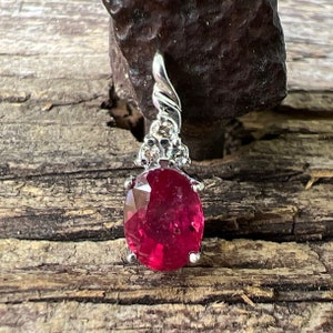 May include: A red gemstone pendant with a silver setting. The gemstone is oval shaped and has a faceted cut. The setting is adorned with smaller white gemstones.