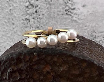 May include: A gold-colored ring with five white pearls. The ring has a unique design with a small, rectangular accent. The pearls are evenly spaced across the top of the ring, creating a delicate and elegant look.