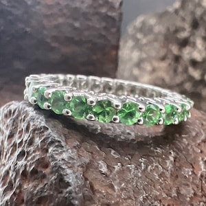 May include: A silver band ring with a row of green gemstones. The ring is set on a rough, dark gray rock.