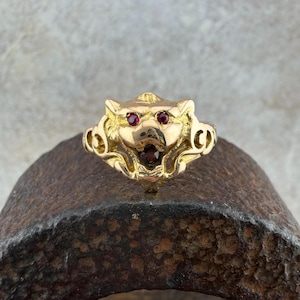 Puede incluir: Un anillo dorado con un diseño de cabeza de animal estilizada, con gemas rojas como ojos y una gema roja oscura en la boca. El anillo presenta detalles intrincados y un estilo vintage, con un diseño único.