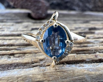 Op de afbeelding: Een gouden ring met een blauwe ovale edelsteen omringd door kleinere heldere edelstenen.