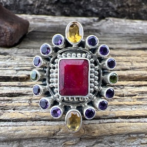 May include: A silver ring with a red ruby gemstone surrounded by a circle of smaller purple and green gemstones.