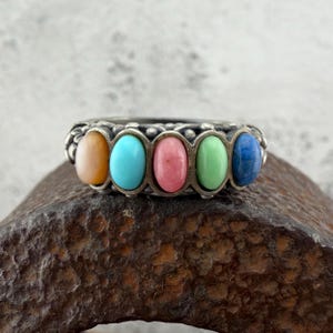 May include: A silver ring featuring five oval gemstones in a row. The gemstones are in shades of orange, turquoise, pink, green, and blue. The ring has a detailed, textured band.