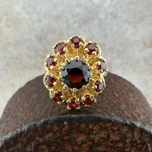 May include: A gold-toned ring featuring a central dark red gemstone surrounded by smaller, matching red gemstones. The ring's design resembles a flower, with the gemstones set in a detailed, ornate setting. The ring is displayed on a textured, brown surface.