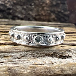 May include: A silver ring with a textured band and a single small, clear gemstone set in the center. The ring is surrounded by small, round silver beads.