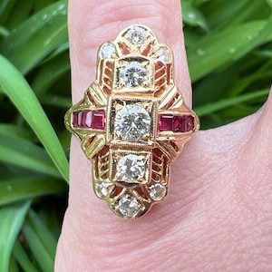 May include: A gold ring with a cross-shaped design featuring a large central diamond and smaller diamonds surrounding it. The ring also has four red ruby stones set in the cross arms.