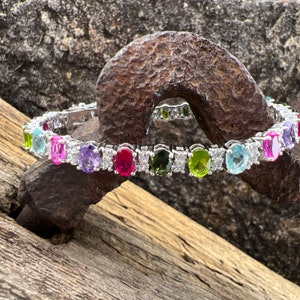 May include: A silver bracelet with oval-shaped gemstones in various colors, including pink, purple, green, and blue. The bracelet is set against a rustic background.