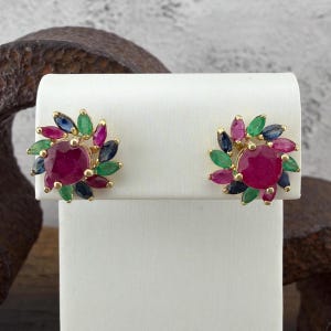 May include: A pair of gold stud earrings. Each earring has a large, round, deep red gemstone at the centre, encircled by smaller, colourful gemstones in green, blue, and pink, arranged in a floral design.
