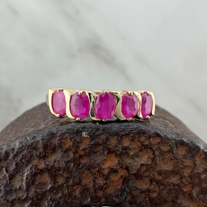 May include: A gold ring featuring five oval-cut vibrant pink gemstones arranged in a row. The stones are set in a polished gold band with curved metal accents separating each individual gemstone, creating a continuous horizontal design.
