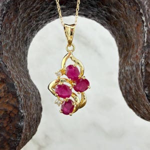 May include: A gold pendant necklace featuring three oval-cut deep pink rubies arranged in a swirling, openwork gold frame. Two small, clear, round-cut accent stones are set within the gold metalwork, which hangs from a delicate gold chain.