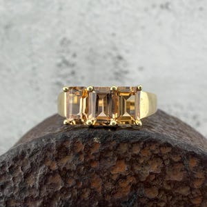 Puede incluir: Un anillo de oro con tres piedras preciosas rectangulares de color marrón claro. El anillo tiene una banda de oro pulido simple. Las piedras están engastadas juntas, creando una línea continua de color.