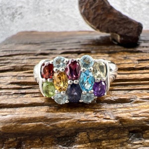 May include: A silver ring with a colorful array of oval-shaped gemstones. The ring features a variety of colors, including red, orange, yellow, green, blue, purple, and clear.