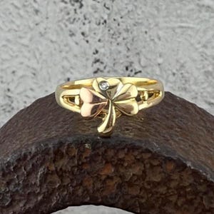 May include: Gold-tone ring featuring a three-leaf clover design with a small diamond accent. The ring has a polished finish and a band with decorative cutouts. The clover has a mix of gold and rose gold tones, creating a two-tone effect.